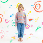 Child wearing a striped shirt and jeans standing against a colorful, child-like drawing background.