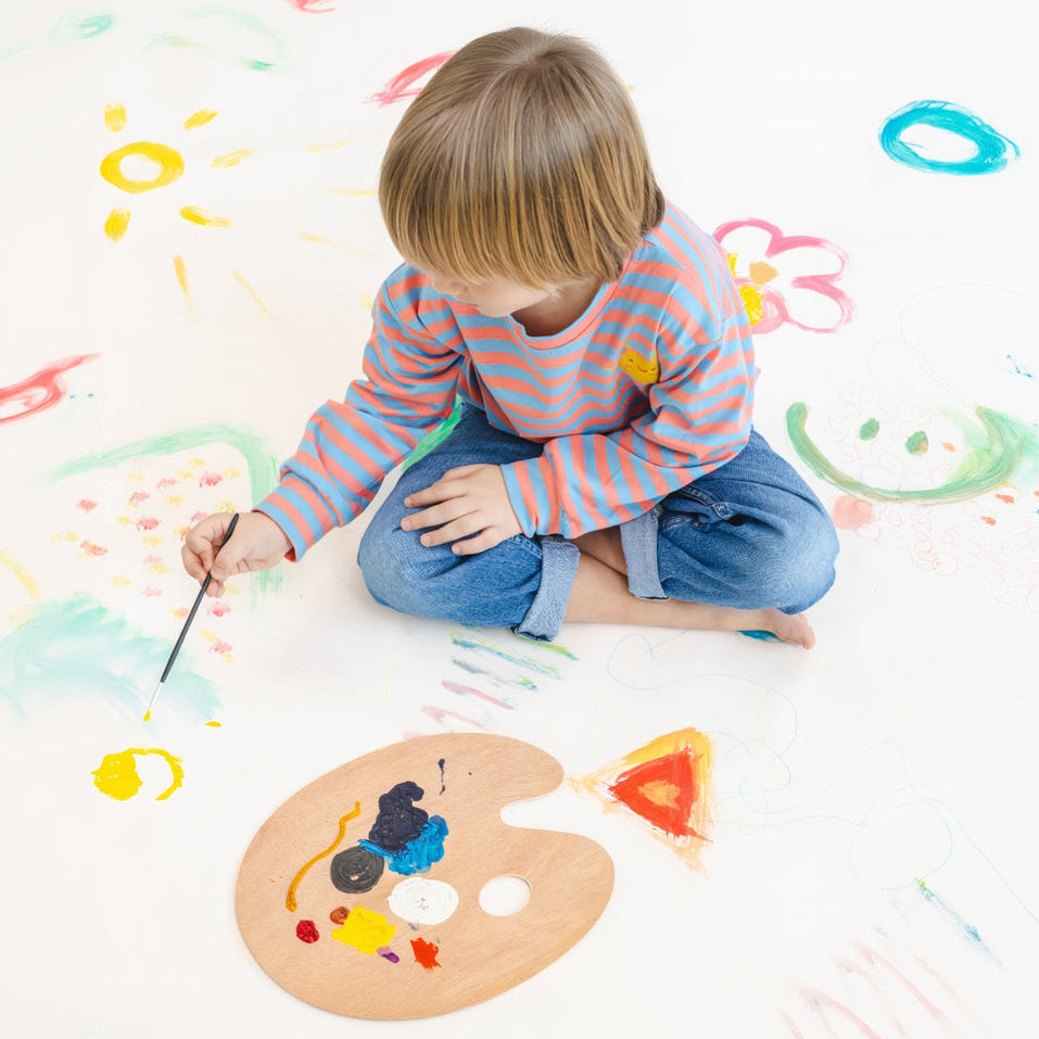 Child sitting on a white floor with colorful paint splashes, holding a paintbrush.