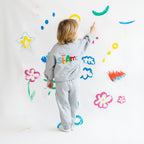 Child in a gray helloshams outfit with colorful text, standing in front of a wall with various child-like drawings.