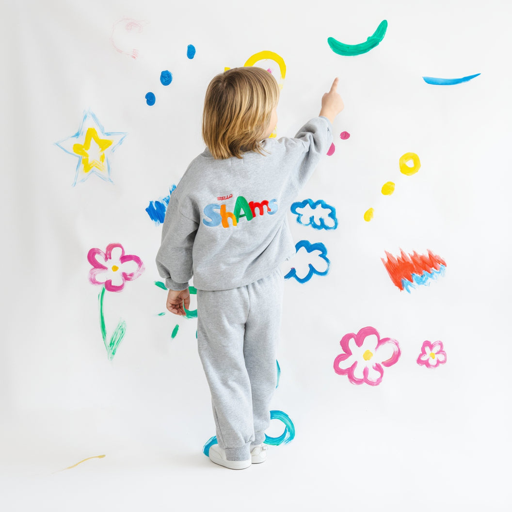 Child in a gray helloshams outfit with colorful text, standing in front of a wall with various child-like drawings.
