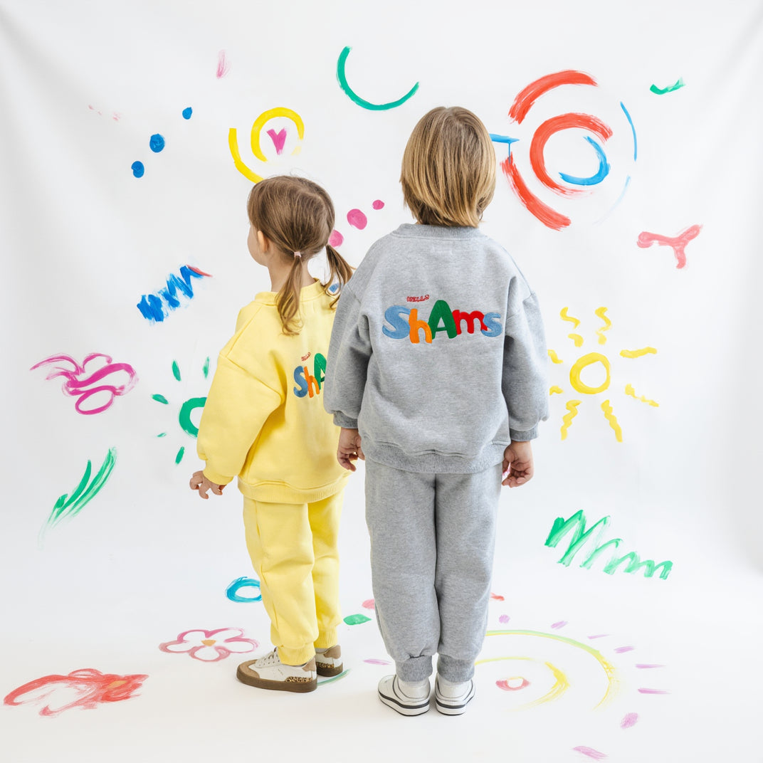 Two children in colorful outfits standing in front of a wall with child-like drawings.