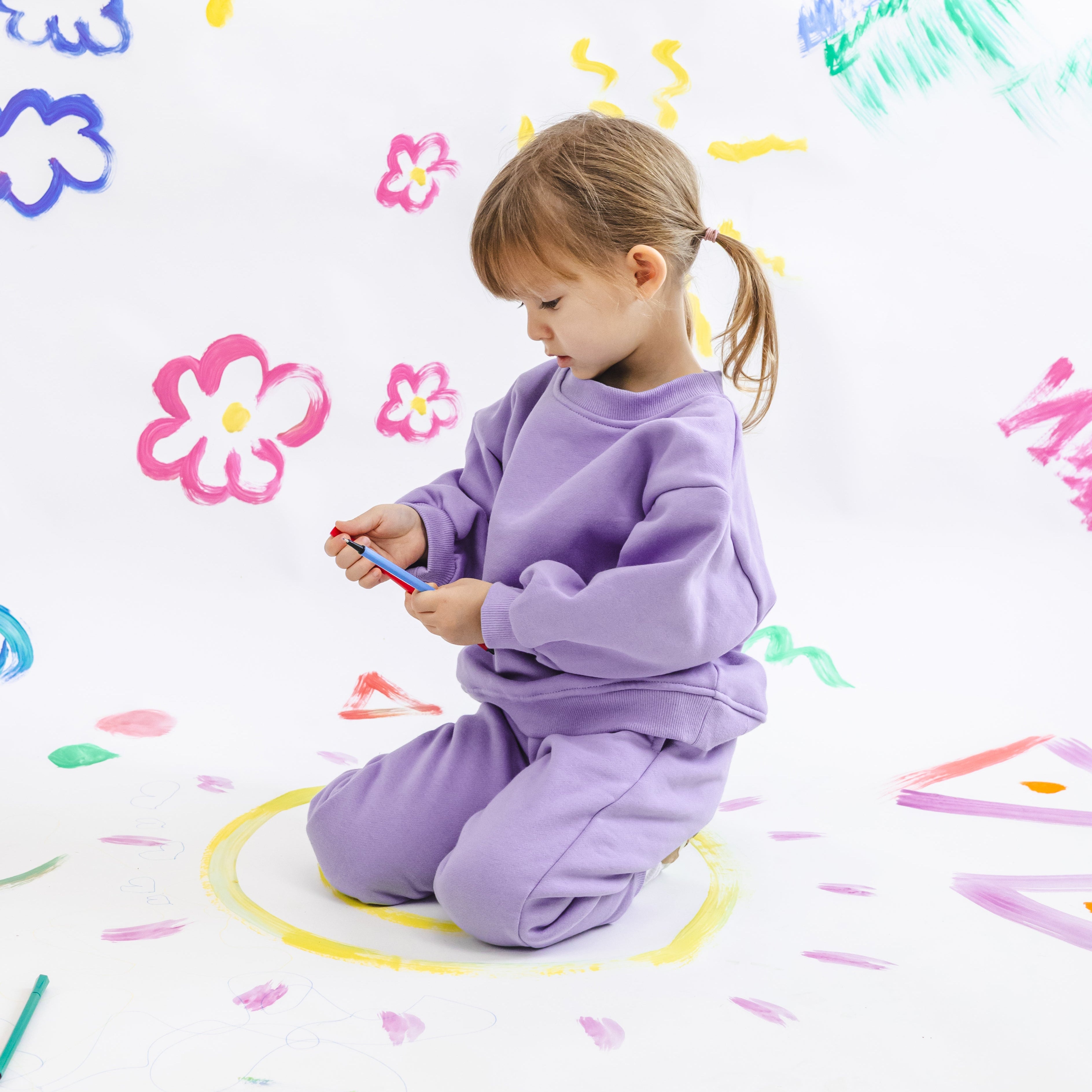 Child in a purple outfit playing with colorful drawings on a white wall.