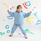 Child in a blue outfit standing smiling in front of a colorful wall with various shapes and designs.