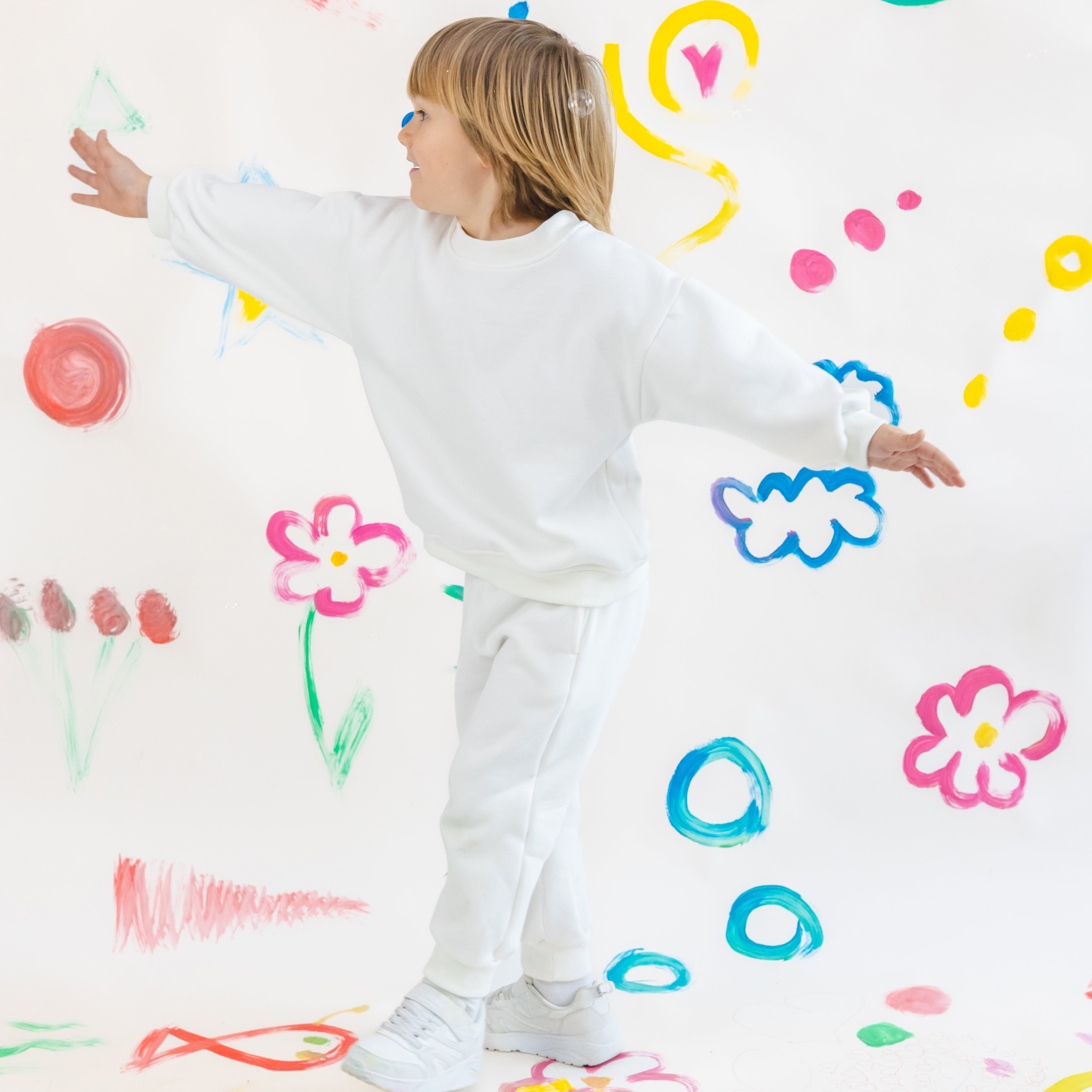 Child in a white outfit standing against a wall with colorful drawings