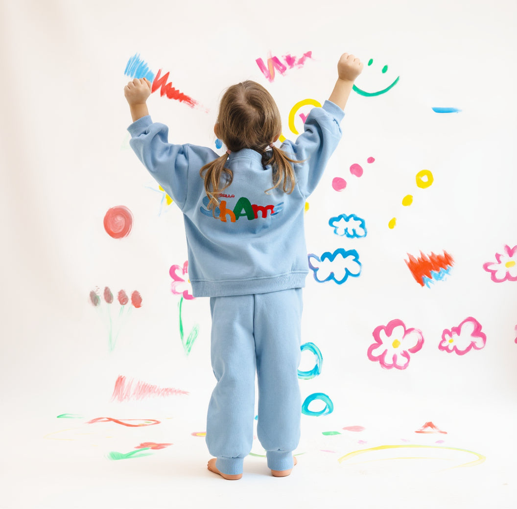 Child wearing a light blue tracksuit with 'Happy' on the back, standing against a colorful, child-like drawing background.