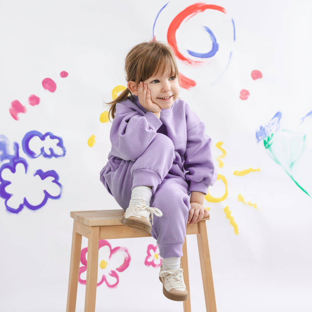 Child in a purple outfit sitting on a wooden stool against a colorful abstract background