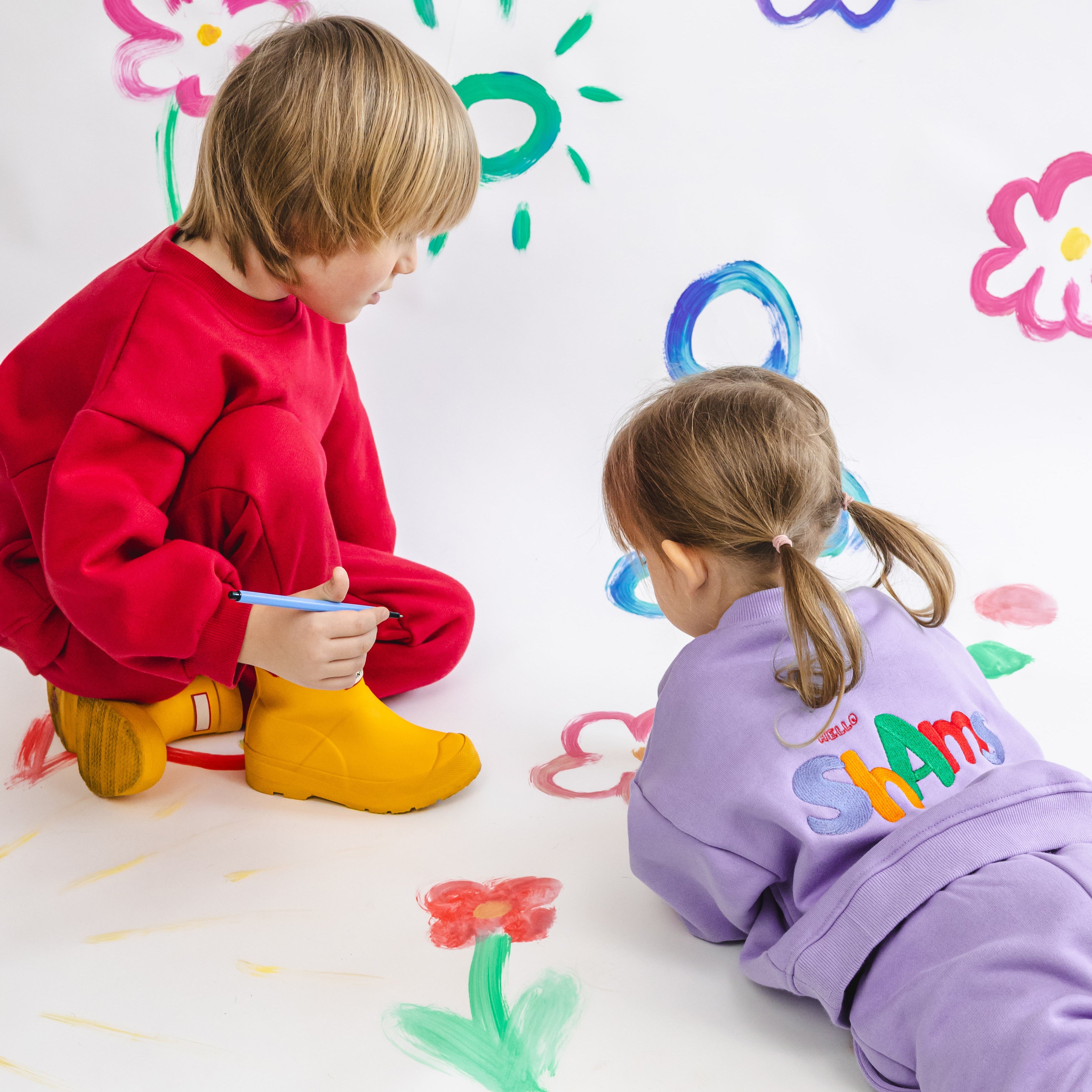 Two children in cozy joggers playing with colorful drawings on a white wall.