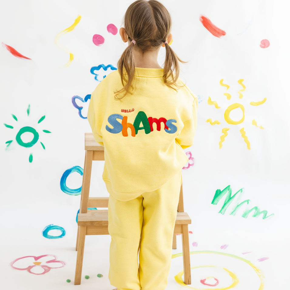 Child wearing a yellow outfit with colorful text, standing on a stool against a wall with painted designs.