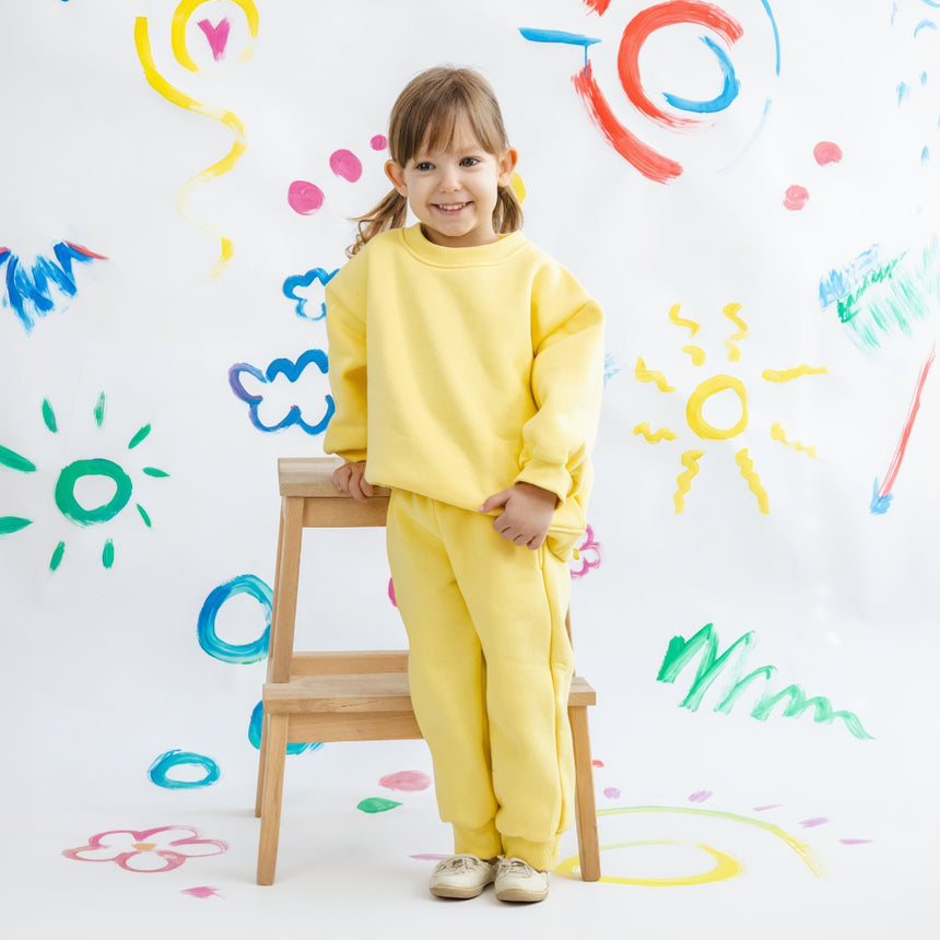 Child in yellow outfit standing on a stool against a wall with colorful child-like drawings.